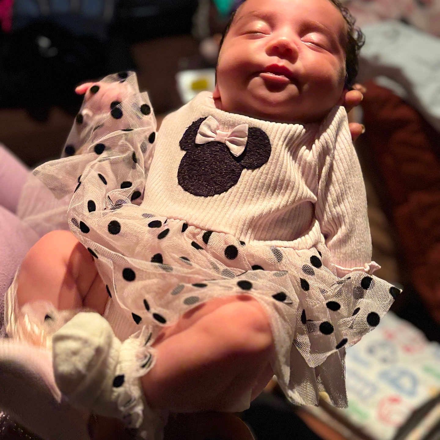 Kailynn is registered to the contest to win money with this photo: newborn, baby, infant, child, portrait, smiling, face, eyes_closed, outfit, dress, polka_dots, bow, mouse_design, held, adult_hands, indoor, bedroom, blanket, soft_lighting, family