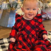Porter is registered to the contest to win money with this photo: baby, child, smiling, red_dress, checkered_pattern, sitting, blanket, holiday, christmas_tree, lights, indoor, cute, festive, happy, person, portrait, floor, decor, ribbon, black_sock