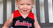 Porter is registered to the contest to win money with this photo: toddler, child, smile, blonde_hair, blue_eyes, cheerleader_outfit, red_clothing, black_clothing, bench, wooden_bench, happy, portrait, sitting, cute, baby, person, indoor, hair_tied, playful, young_child