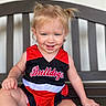 toddler, child, smile, blonde_hair, blue_eyes, cheerleader_outfit, red_clothing, black_clothing, bench, wooden_bench, happy, portrait, sitting, cute, baby, person, indoor, hair_tied, playful, young_child