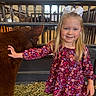 child, girl, calf, animal, barn, hay, smile, bow, blonde_hair, long_sleeves, purple_dress, indoor, livestock, petting, fence, young, cute, friendly, farm, happy