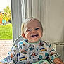 toddler, smiling, baby, high_chair, bib, indoor, window, curtains, construction_vehicles, happy, child, face, teeth, clothing, person, furniture, home, daylight, greenery, background