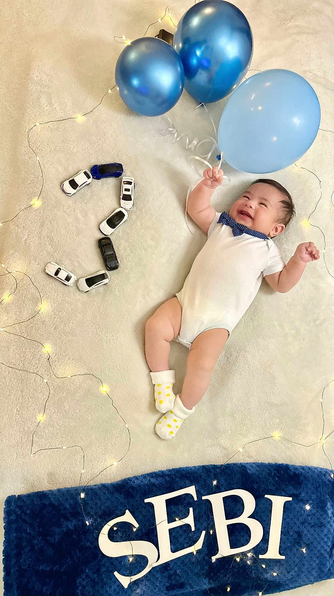 Elian Sebastian is registered to the contest to win money with this photo: baby, balloons, birthday, blanket, blue, celebration, child, cute, decorations, happy, indoor, infant, lights, number_two, playful, polka_dot_socks, smiling, soft_texture, toy_cars, white_onesie