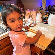 Isabella is registered to the contest to win money with this photo: child, girl, kitchen, rolling_pin, dough, countertop, wooden_cabinets, flour, utensils, canisters, pink_shirt, curly_hair, baking, cooking, home, appliance, stove, sink, expression, portrait
