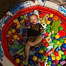 Lucas is registered to the contest to win money with this photo: toddler, child, inflatable_pool, plastic_balls, wooden_blocks, toy, indoor, carpet, colorful, playtime, cute, person, smiling, ball, green_toy, orange_ball, purple_ball, yellow_ball, red_ball, casual_clothing