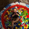 toddler, child, ball_pit, colorful_balls, wooden_blocks, indoor, carpet, toy, playtime, inflatable, person, smiling, cute, casual_clothing, green_plush_toy, basketball_toy, soccer_ball_toy, relaxing, bright_colors, happy