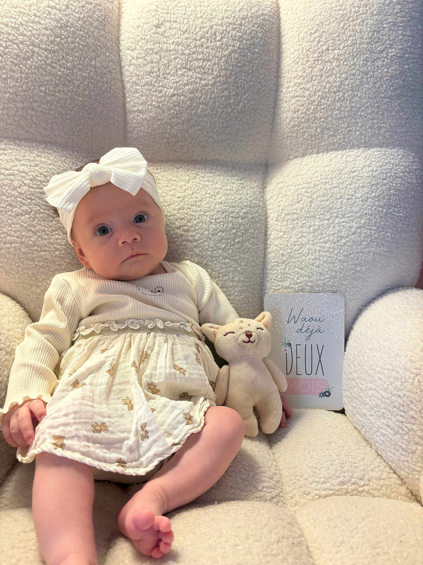 Isaé a rejoint le concours — aidez-le/la à gagner de superbes lots ! baby, infant, headband, white_bow, dress, skirt, stuffed_animal, plush_toy, milestone_card, armchair, textured_fabric, portrait, indoor, cute, bare_feet, toes, wide_eyes, soft_light, neutral_colors, sitting