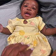Klarity joined the competition — help win amazing prizes! baby, child, smiling, yellow_dress, fruit_pattern, bed, gray_sheets, hand, lying_down, cute, happy, infant, person, portrait, skin, curly_hair, indoors, fabric, clothing, playful