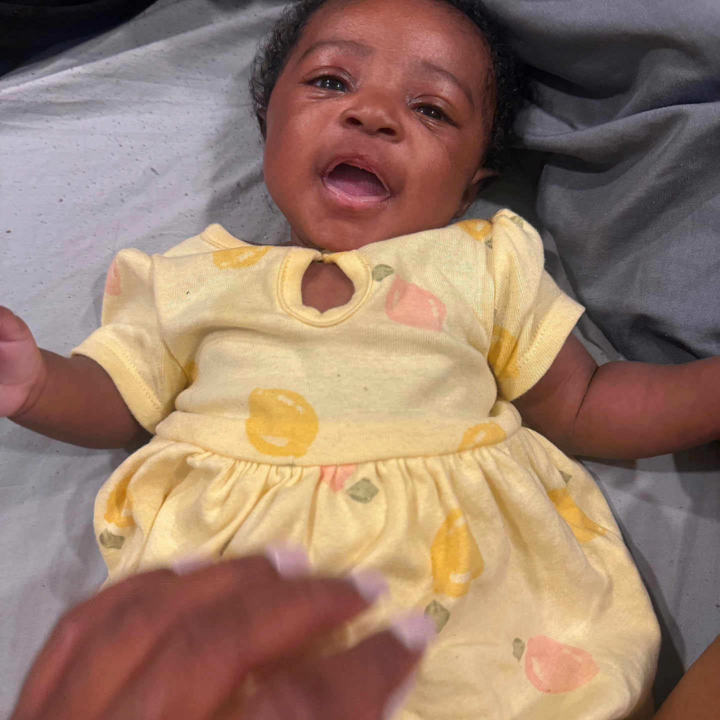 Klarity joined the competition — help win amazing prizes! baby, child, smiling, yellow_dress, fruit_pattern, bed, gray_sheets, hand, lying_down, cute, happy, infant, person, portrait, skin, curly_hair, indoors, fabric, clothing, playful