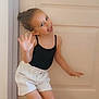 child, girl, smiling, happy, pose, indoor, door, shorts, black_top, white_shorts, hair_bun, waving_hand, playful, floor_tiles, casual_clothing, youth, cute, person, expression, fun