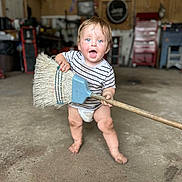 Luis is registered to the contest to win money with this photo: baby, child, toddler, standing, smiling, surprised, blue_eyes, broom, cleaning, garage, workshop, 工具, striped_shirt, diaper, barefoot, indoor, portrait, floor, tools, funny