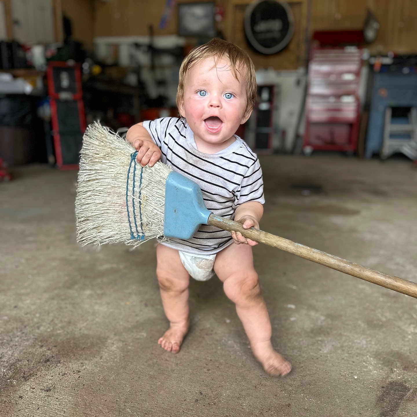 Luis is registered to the contest to win money with this photo: baby, child, toddler, standing, smiling, surprised, blue_eyes, broom, cleaning, garage, workshop, 工具, striped_shirt, diaper, barefoot, indoor, portrait, floor, tools, funny