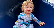 Luis is registered to the contest to win money with this photo: child, toddler, playground, slide, blue_slide, sitting, outdoor, portrait, blond_hair, blue_eyes, pajamas, clothing, shoes, smiling, face, play_area, wood_chips, sky, sunlight, playtime