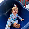 child, toddler, playground, slide, blue_slide, sitting, outdoor, portrait, blond_hair, blue_eyes, pajamas, clothing, shoes, smiling, face, play_area, wood_chips, sky, sunlight, playtime