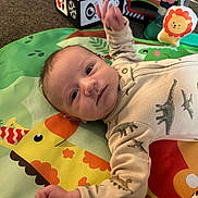 Dayton is registered to the contest to win money with this photo: baby, infant, play_mat, dinosaur_onesie, animal_print, colorful, cute, child, indoor, soft_toys, smiling, hand_raised, portrait, young_child, floor_play, clothing, face, head, person, newborn