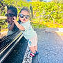 toddler, child, sunglasses, reflection, car, parking_lot, outdoor, sunlight, greenery, pavement, clothing, shorts, sandals, cute, portrait, happy, person, summer, casual, fashion