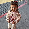 child, toddler, smiling, outdoor, sidewalk, concrete, pigtails, red_bows, cards, street, curb, casual_clothing, sweatshirt, pants, sneakers, sunlight, happy, young_child, person, standing
