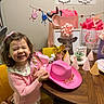 child, girl, smiling, pink_clothing, birthday, party_hat, cowboy_hat, sash, gift_bags, table, chair, decorations, birthday_party, happy, indoor, celebration, present, wall_art, wooden_table, yellow_chair