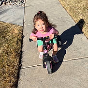 Emberlyn is registered to the contest to win money with this photo: child, bike, sidewalk, outdoor, sunny, shadow, grass, rocks, pink_shirt, green_shorts, sneakers, playful, young_child, concrete, daylight, hair_tied, small_bike, riding, happy, person