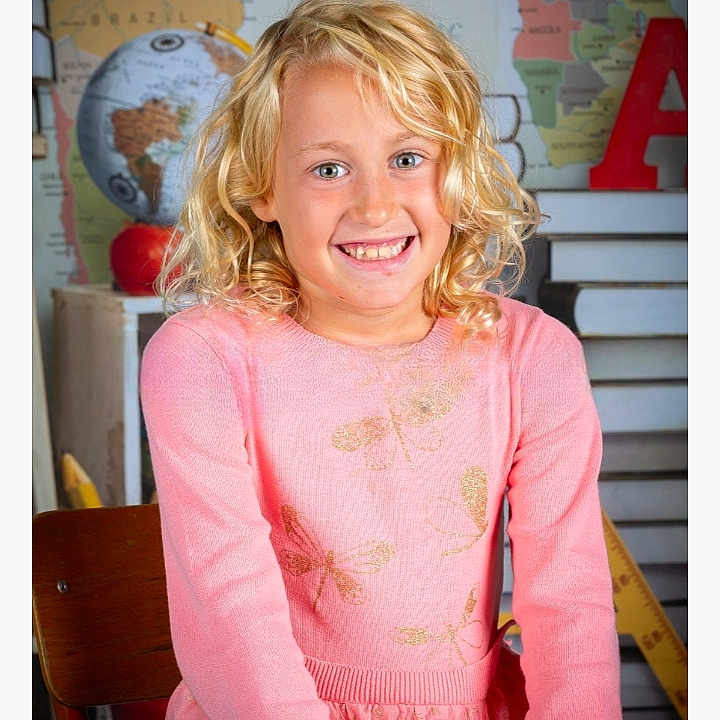 Lana a rejoint le concours — aidez-le/la à gagner de superbes lots ! background, blonde_hair, casual_clothing, chair, child, classroom, curly_hair, cute, dragonfly_pattern, education, girl, globe, happy, indoor, map, pink_sweater, portrait, seated, smiling, young
