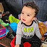 Axel is registered to the contest to win money with this photo: toddler, child, curly_hair, sitting, car_interior, toys, clothing, shirt, text_on_clothing, footwear, curious, drool, messy_background, box, plastic_container, stuffed_toy, car_seat, person, portrait, indoors
