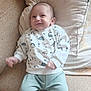 baby, infant, smiling, sweater, dinosaur_pattern, pants, couch, pillow, indoor, child, happy, cute, portrait, seated, comfort, young_child, clothing, face, person, home