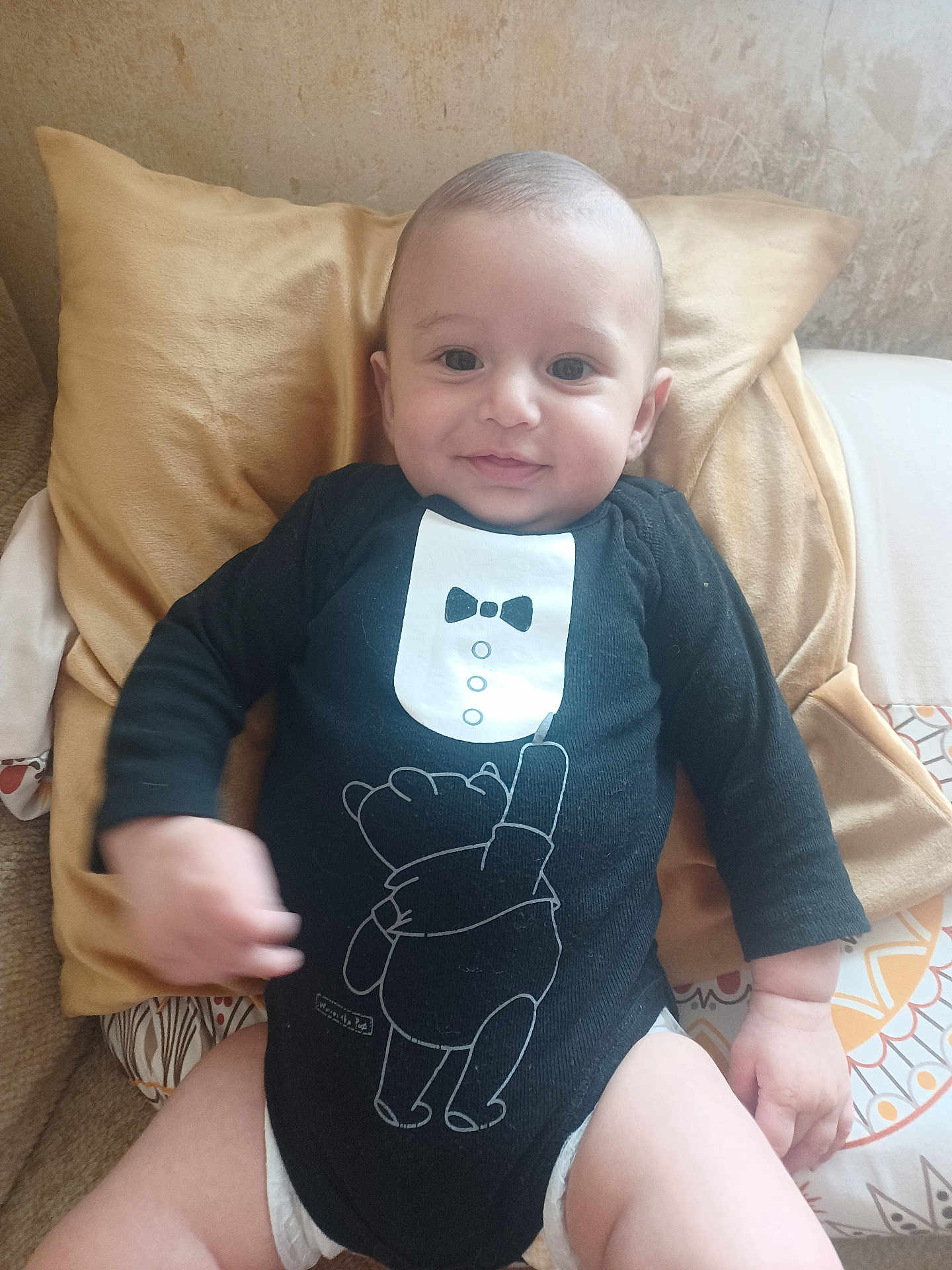 Lowen participe au concours pour gagner de l'argent avec cette photo : baby, infant, smile, black_onesie, tuxedo_print, bear_print, pillow, bed, blanket, hand, face, cute, child, indoor, person, sitting, happy, diaper, couch, patterned_bedding