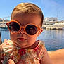 baby, sunglasses, child, floral_dress, water, outdoor, sunlight, person, hand, summer, cute, portrait, fashion, head, face, skin, expression, bright, vacation, holding