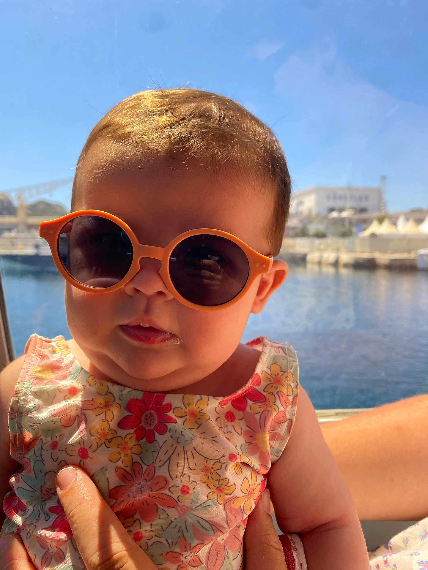 Emy participe au concours pour gagner de l'argent avec cette photo : baby, sunglasses, child, floral_dress, water, outdoor, sunlight, person, hand, summer, cute, portrait, fashion, head, face, skin, expression, bright, vacation, holding
