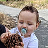 Ewan is registered to the contest to win money with this photo: baby, child, toddler, outdoor, portrait, pine_cone, pacifier, white_shirt, brown_hair, face, hands, sitting, adult, arm, sidewalk, path, grass, trees, nature, daylight