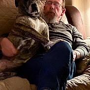 Bella is registered to the contest to win money with this photo: dog, person, indoor, couch, glasses, sitting, blanket, curtains, relaxed, pet, lap, long_sleeve_shirt, casual_clothing, footwear, red_shoes, brown, white, furniture, home, beard
