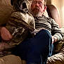 dog, person, indoor, couch, glasses, sitting, blanket, curtains, relaxed, pet, lap, long_sleeve_shirt, casual_clothing, footwear, red_shoes, brown, white, furniture, home, beard