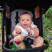 Saige joined the competition — help win amazing prizes! baby, stroller, child, outdoor, grass, greenery, bottle, playful, smiling, tongue_out, clothing, footwear, seatbelt, toy, happy, person, infant, nature, sitting, cute