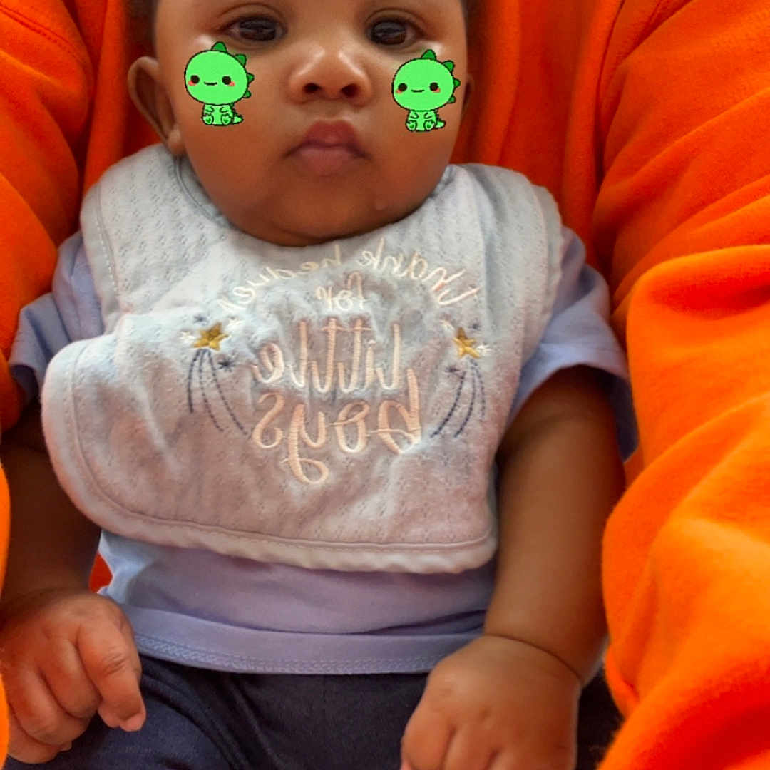 Kaison is registered to the contest to win money with this photo: baby, child, infant, bib, clothing, orange_sweater, sitting, cute, face_filter, digital_sticker, green_dinosaur, hands, legs, socks, portrait, indoor, person, holding, expressionless, casual