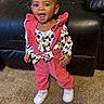 toddler, child, smiling, standing, pink_overalls, patterned_shirt, white_shoes, carpet, leather_couch, indoor, happy, portrait, earring, clothing, person, baby, cute, fashion, home, young_child