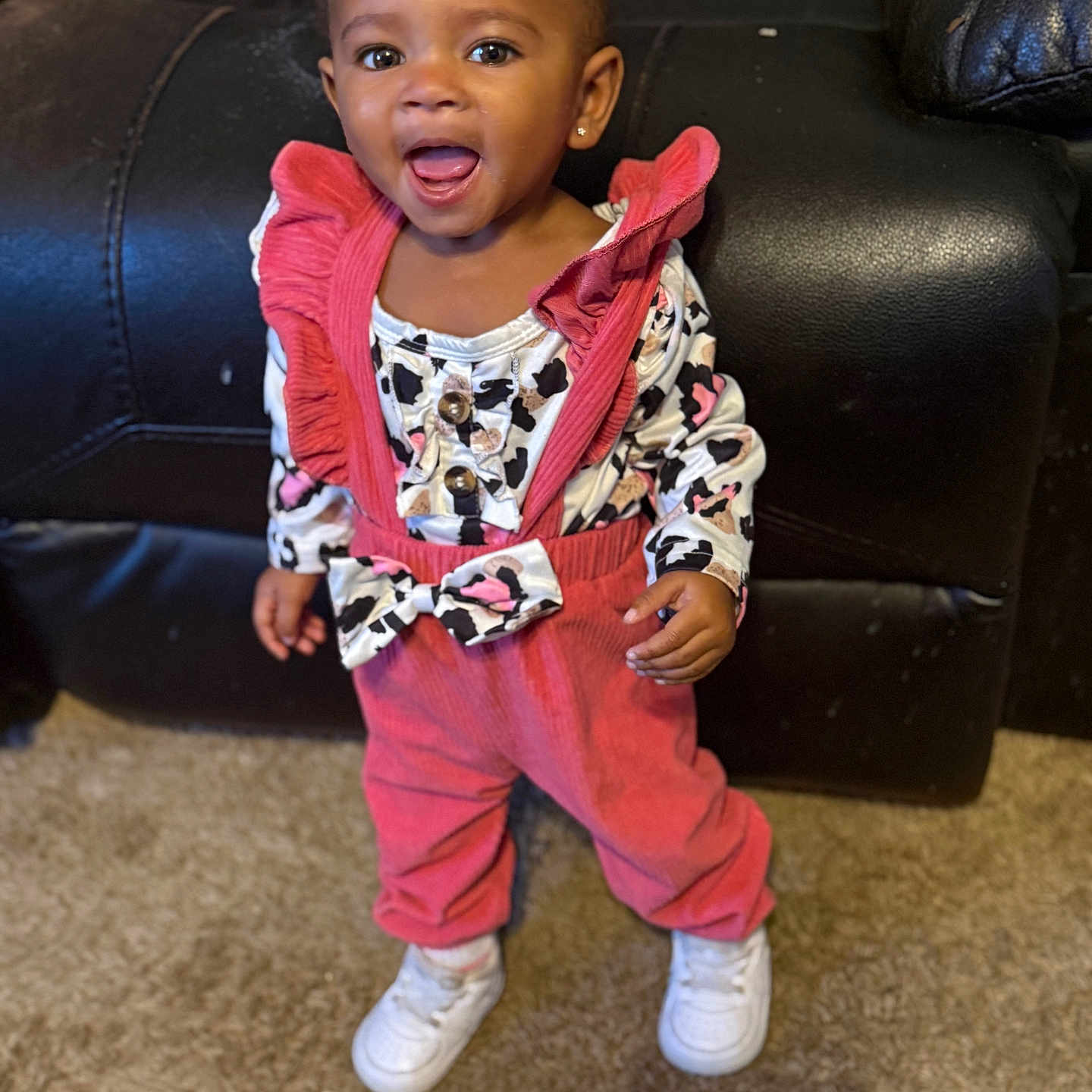 Aubrey joined the competition — help win amazing prizes! toddler, child, smiling, standing, pink_overalls, patterned_shirt, white_shoes, carpet, leather_couch, indoor, happy, portrait, earring, clothing, person, baby, cute, fashion, home, young_child