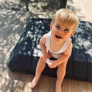 Malo participe au concours pour gagner de l'argent avec cette photo : background_blur, barefoot, blonde_hair, child, cushion, cute, daylight, face, happy, nature, outdoor, person, shadow, sitting, small_object, smiling, sunlight, toddler, white_clothing, wood_floor