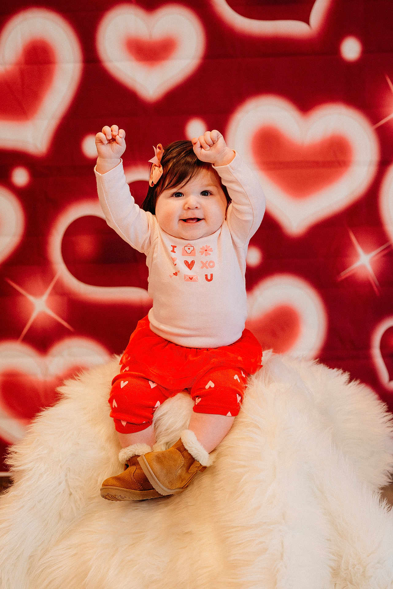 Mia is registered to the contest to win money with this photo: baby, child, smiling, raised_hands, pink_bow, white_shirt, red_skirt, heart_pattern, background, fluffy_surface, boots, cute, portrait, indoor, happy, sitting, warm_clothing, celebration, festive, joyful