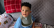 Weston is registered to the contest to win money with this photo: child, curly_hair, blue_outfit, bib, blanket, couch, home_interior, wall_art, decor, plush_blanket, window_blinds, red_blanket, patterned_blanket, sitting, indoors, person, smile, face, cozy, relaxing