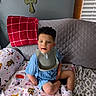 child, curly_hair, blue_outfit, bib, blanket, couch, home_interior, wall_art, decor, plush_blanket, window_blinds, red_blanket, patterned_blanket, sitting, indoors, person, smile, face, cozy, relaxing