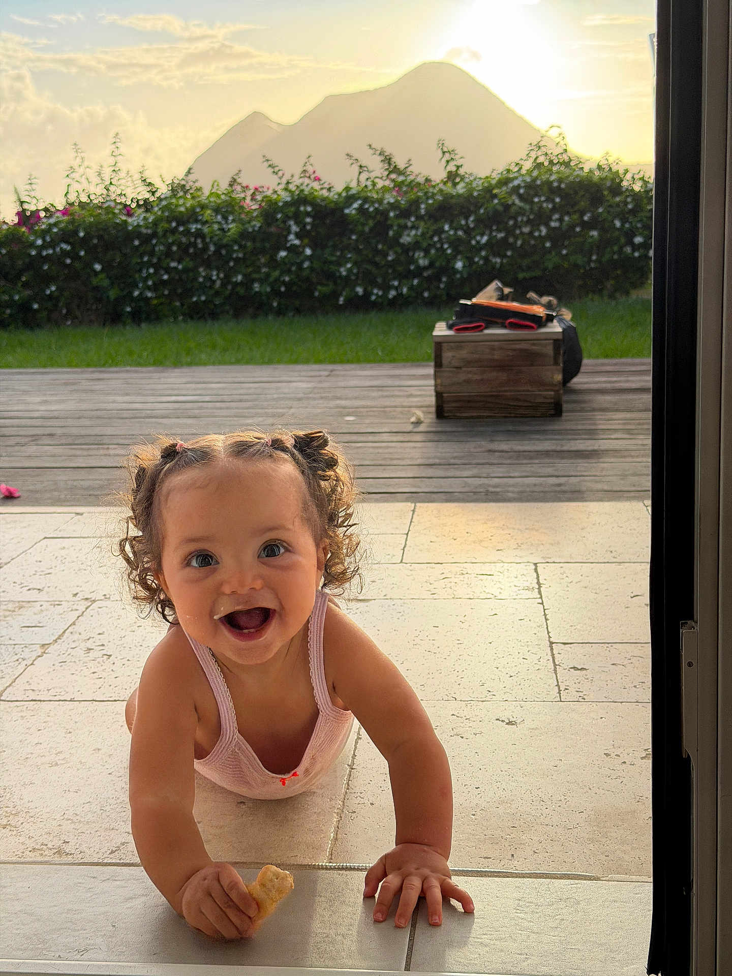 Mahéa a rejoint le concours — aidez-le/la à gagner de superbes lots ! toddler, child, crawling, curly_hair, pigtails, snack, smiling, outdoor, garden, grass, mountains, sunlight, tile_floor, happy, person, nature, daylight, playful, young_child, curiosity