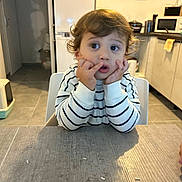 Mylann participe au concours pour gagner de l'argent avec cette photo : toddler, child, curly_hair, striped_sweater, table, kitchen, indoor, face, hands, expression, rice_grains, white_chair, floor_tiles, cabinet, microwave, appliance, curious, sitting, young_child, person