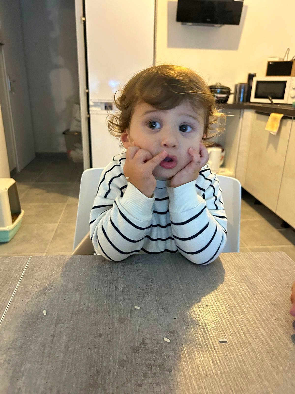 Mylann participe au concours pour gagner de l'argent avec cette photo : toddler, child, curly_hair, striped_sweater, table, kitchen, indoor, face, hands, expression, rice_grains, white_chair, floor_tiles, cabinet, microwave, appliance, curious, sitting, young_child, person