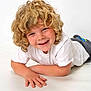 Ashton is registered to the contest to win money with this photo: child, curly_hair, smiling, white_tshirt, pants, sneakers, lying_down, studio, portrait, happy, boy, casual_clothing, playful, young, innocent, cute, light_background, human, face, hands