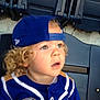 child, toddler, curly_hair, baseball_cap, blue_cap, sports_jersey, stadium_seat, looking_up, outdoor, young_child, casual_clothing, blond_hair, portrait, seat_back, curious_expression, daylight, person, hat, captured_moment, baseball