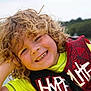 Ashton is registered to the contest to win money with this photo: child, smiling, curly_hair, life_jacket, yellow_shirt, outdoor, lake, water, forest, summer, happy, portrait, young, person, nature, daylight, casual_clothing, recreation, fun, closeup