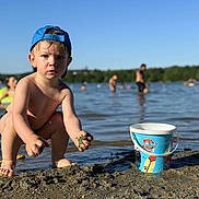 Aydenn a rejoint le concours — aidez-le/la à gagner de superbes lots ! toddler, child, blue_cap, sand, mud, bucket, paw_patrol, water, lake, outdoor, summer, playing, barefoot, wet, people, background, nature, sunny, recreation, reflection