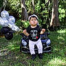 toddler, child, boy, outdoor, grass, trees, toy_car, balloons, black_shirt, white_pants, hat, cap, nature, sunlight, greenery, portrait, standing, cute, fashion, playtime