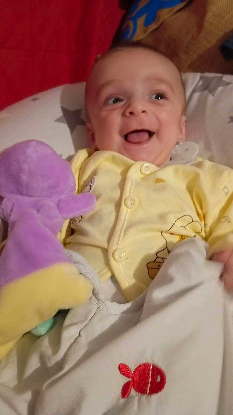 Eden a rejoint le concours — aidez-le/la à gagner de superbes lots ! baby, smiling, yellow_onesie, stuffed_toy, purple_toy, blanket, red_fish, happy, infant, portrait, child, cute, indoor, cozy, soft_toy, bedtime, face, person, playful, comfort