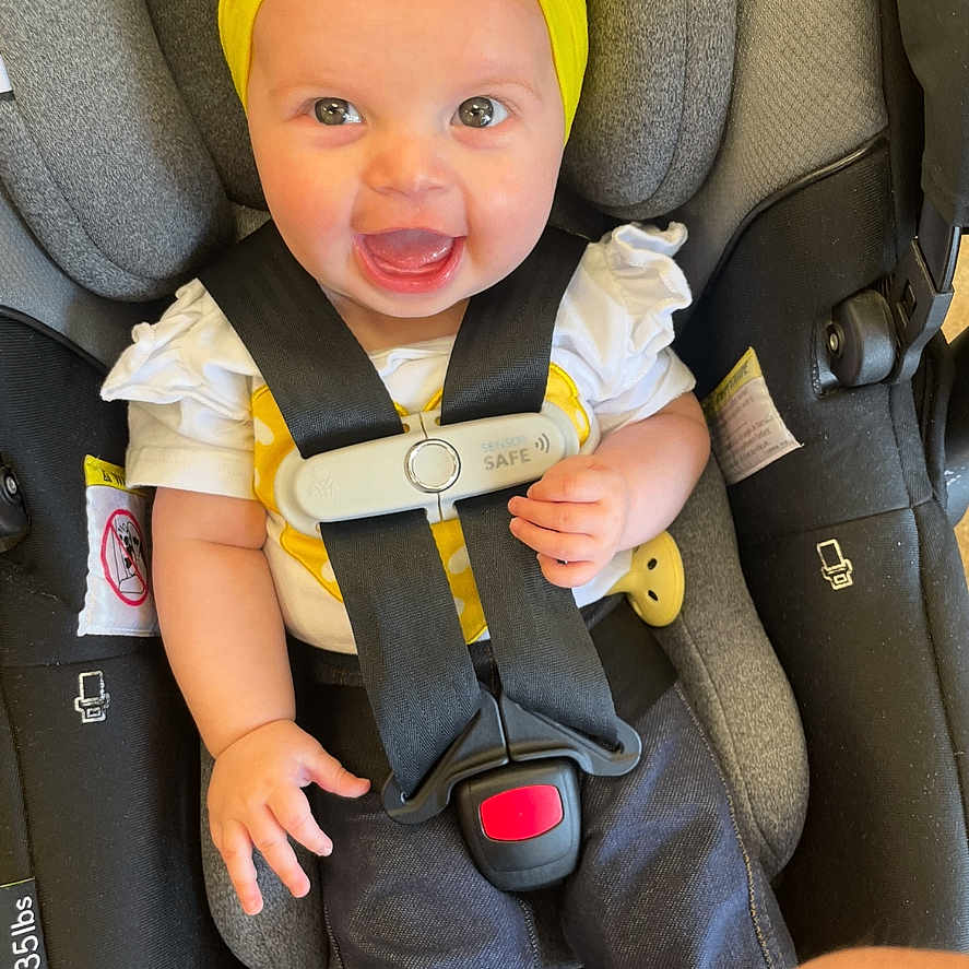 Kali is registered to the contest to win money with this photo: baby, car_seat, yellow_headband, smiling, child, infant, straps, happy, sitting, clothing, seatbelt, cute, portrait, young_child, indoor, person, headwear, bright_colors, security, car_interior
